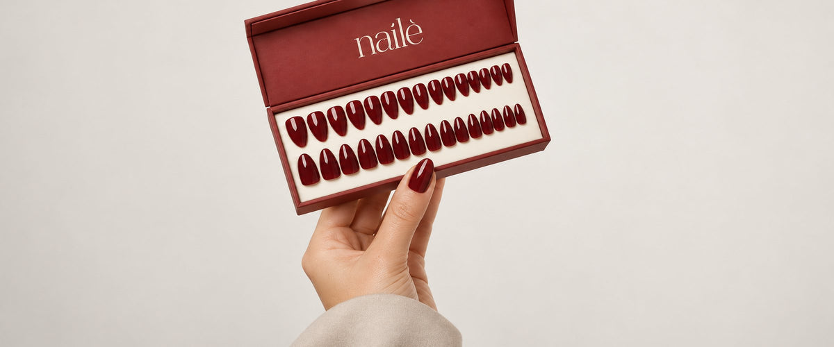 A hand holding an open Nailé Creations box of deep burgundy press-on nails