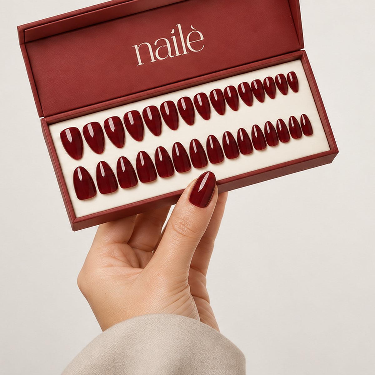 A hand holding an open Nailé Creations box of deep burgundy press-on nails